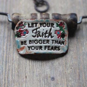 May include: A rectangular pendant with the words "LET YOUR Faith BE BIGGER THAN YOUR FEARS" in black lettering. The pendant has a floral design with red and blue flowers. The pendant is attached to a metal and wood handle.
