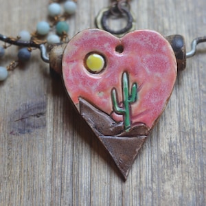 May include: A heart-shaped ceramic pendant featuring a pink and orange sunset sky, a yellow sun, a green saguaro cactus, and brown desert terrain. The pendant is attached to a dark metal wire necklace adorned with small, round, light blue beads.
