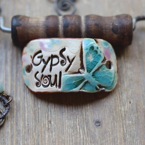 May include: A ceramic pendant necklace with a butterfly design and the words "Gypsy Soul" on a white background. The necklace is made with a brown wooden toggle clasp and a chain with light blue and green beads.