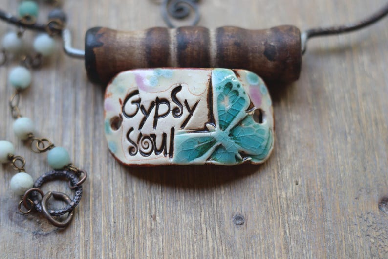 May include: A ceramic pendant necklace with a butterfly design and the words "Gypsy Soul" on a white background. The necklace is made with a brown wooden toggle clasp and a chain with light blue and green beads.