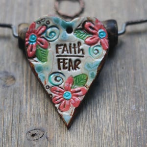 May include: A heart-shaped ceramic pendant featuring the embossed text FAITH FEAR in black lettering. The surface is decorated with raised red flowers, green leaves, and blue swirls on a cream background, suspended from a dark metal wire necklace.