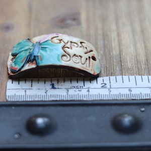 May include: A ceramic charm with a butterfly design and the words "Gypsy Soul" in a white glaze. The charm is about 2 inches long.
