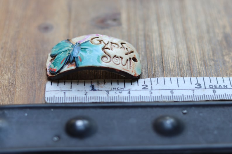 May include: A ceramic charm with a butterfly design and the words "Gypsy Soul" in a white glaze. The charm is about 2 inches long.