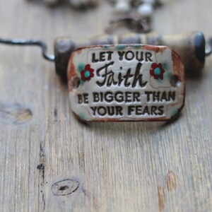 May include: A rectangular ceramic pendant with rounded edges featuring the embossed black text LET YOUR FAITH BE BIGGER THAN YOUR FEARS. The off-white surface is accented with small red and turquoise floral details and a rustic brown border.