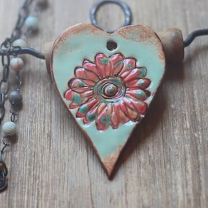 May include: A ceramic heart pendant necklace with a floral design. The heart is painted in a light blue glaze with a pink and green flower in the center. The necklace is made with a black chain and a wooden bead.
