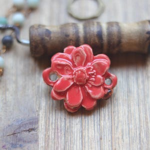 May include: A bright coral-red ceramic flower bead with a glossy finish and two side holes for threading. The flower features layered petals and a textured center, positioned near a wooden handle and a strand of light blue and grey beads.