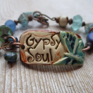 May include: A bracelet with a ceramic charm that says "Gypsy Soul" and features a blue butterfly design. The bracelet is made with copper wire, blue and green glass beads, and a white bead.