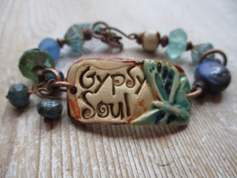 May include: A bracelet with a ceramic charm that says "Gypsy Soul" and features a blue butterfly design. The bracelet is made with copper wire, blue and green glass beads, and a white bead.