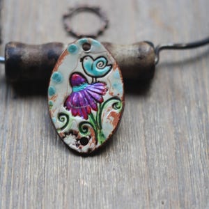 May include: Oval pendant necklace with a hand-painted design of a purple flower and a blue bird. The pendant is on a dark metal chain. The background is a wooden surface.