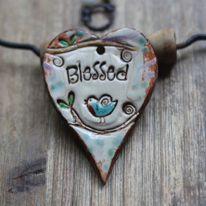 May include: A heart-shaped ceramic pendant with the word "Blessed" inscribed. The pendant features a small bird illustration and decorative floral accents in blue, red, and green. It hangs from a black wire and wooden handle.