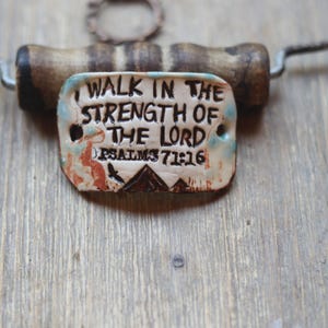 May include: A handmade ceramic pendant with the inscription "I WALK IN THE STRENGTH OF THE LORD PSALMS 71:16". The pendant has a rustic design with brown, blue, and orange accents. It is attached to a dark metal wire and a wooden handle.