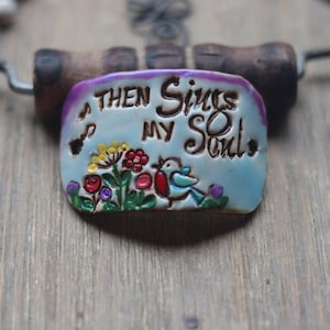 May include: A light blue ceramic pendant with the text "Then Sings My Soul" in white lettering. The pendant is decorated with a small bird and flowers in red, yellow, and green.