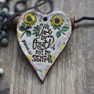 May include: A white ceramic heart-shaped pendant with a floral design and the text "We live by faith, not by sight" and the scripture reference "2 Corinthians 5:7".