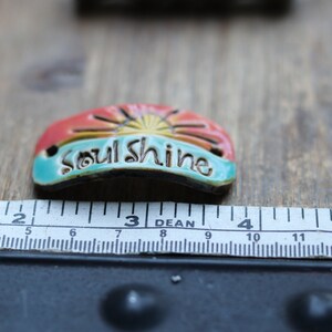 Puede incluir: Colgante de cer&aacute;mica con la inscripci&oacute;n "soul shine" en una pancarta. El colgante mide aproximadamente 5 cm de largo y presenta un dise&ntilde;o de amanecer con tonos rojos, naranjas y amarillos. La pancarta es turquesa.