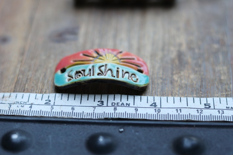 Puede incluir: Colgante de cer&aacute;mica con la inscripci&oacute;n "soul shine" en una pancarta. El colgante mide aproximadamente 5 cm de largo y presenta un dise&ntilde;o de amanecer con tonos rojos, naranjas y amarillos. La pancarta es turquesa.
