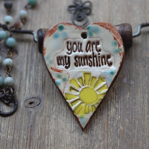 May include: A heart-shaped pendant with the text "you are my sunshine" and a yellow sun design. The pendant is white with brown and blue accents. A beaded necklace and a metal wire are also visible.