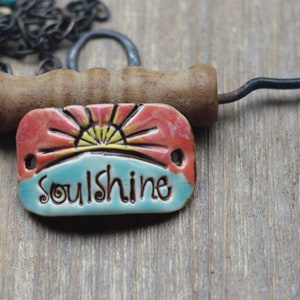 Puede incluir: Colgante de cer&aacute;mica con la palabra "Soulshine" en una fuente caprichosa. El colgante presenta un dise&ntilde;o de amanecer en rojo, amarillo y azul. Un mango de madera y una cadena de metal son visibles en el fondo.
