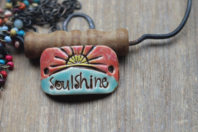 Puede incluir: Colgante de cer&aacute;mica con la palabra "Soulshine" en una fuente caprichosa. El colgante presenta un dise&ntilde;o de amanecer en rojo, amarillo y azul. Un mango de madera y una cadena de metal son visibles en el fondo.