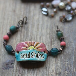 Puede incluir: Una pulsera hecha a mano con una pieza central de cer&aacute;mica que presenta un dise&ntilde;o de amanecer y la palabra "soulshine". La pulsera incluye cuentas turquesas, coral y verdes, sobre un fondo de madera.