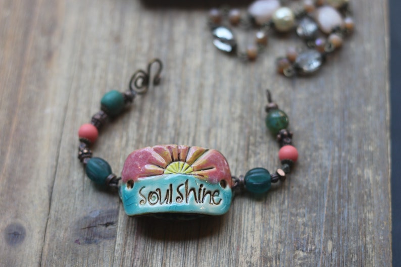 Puede incluir: Una pulsera hecha a mano con una pieza central de cer&aacute;mica que presenta un dise&ntilde;o de amanecer y la palabra "soulshine". La pulsera incluye cuentas turquesas, coral y verdes, sobre un fondo de madera.