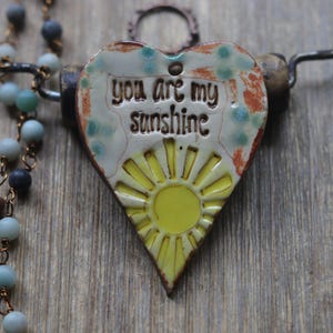 May include: A heart-shaped ceramic pendant featuring a yellow sun graphic and the text "you are my sunshine" in dark lettering. The pendant has a cream-colored background with blue and orange accents, suspended on a wire necklace with blue beads.