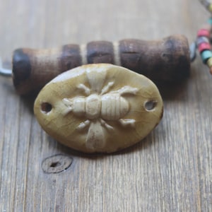 May include: A light beige, oval-shaped ceramic bead with a bee design, featuring two holes for stringing. A wooden handle and a beaded necklace are in the background, all resting on a wooden surface.