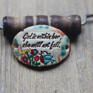 May include: A white oval ceramic pendant with the text "God is within her, she will not fall." The pendant is decorated with colorful flowers and a blue bird.