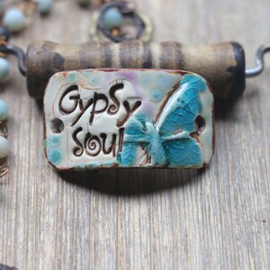 May include: A rectangular ceramic pendant featuring the text "Gypsy Soul" in black lettering alongside a blue butterfly graphic with a crackled glaze finish. The pendant is attached to a beaded necklace with light blue and grey round beads.
