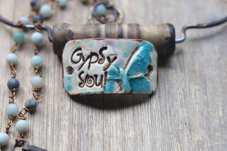 May include: A rectangular ceramic pendant featuring the text "Gypsy Soul" in black lettering alongside a blue butterfly graphic with a crackled glaze finish. The pendant is attached to a beaded necklace with light blue and grey round beads.