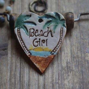May include: A heart-shaped ceramic pendant featuring a hand-painted beach scene with green palm trees, a yellow sun, blue water, and a textured brown sand base. The text Beach Girl is inscribed in the center. It hangs from a dark metal wire.