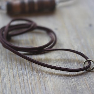 May include: A close-up of a dark brown leather cord with a small silver jump ring. The cord is lying on a wooden surface.