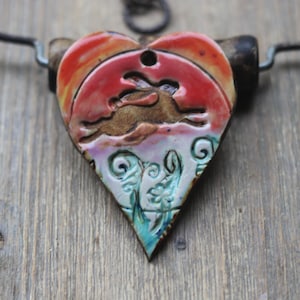 May include: Heart-shaped pendant necklace with a running hare design. The pendant features a red and orange sky, a brown hare, and a turquoise and white floral design. The necklace is on a wooden surface.