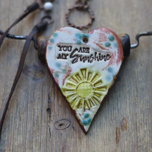 May include: A heart-shaped ceramic pendant featuring a yellow sun relief and the embossed text "YOU ARE MY SUNSHINE" in black. The pendant has a cream-colored base with blue and reddish-brown accents, suspended from a dark brown leather cord.