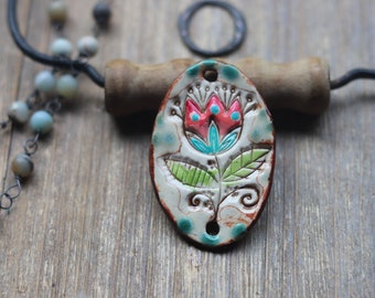 May include: Oval ceramic pendant with a floral design. The pendant features a red and turquoise flower with green leaves on a cream background. The pendant is accented with turquoise dots and a brown border. A beaded necklace and a metal ring are in the background.