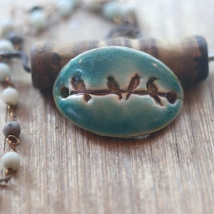 May include: An oval ceramic pendant with a teal crackle glaze featuring a horizontal line of four small birds etched into the surface. The pendant is accompanied by a beaded necklace strand with light blue and grey round beads on a dark wire.