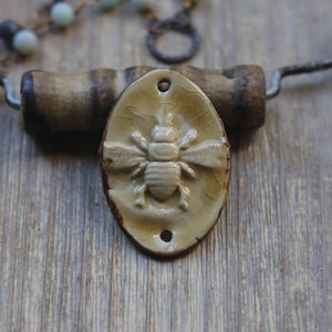 May include: An oval-shaped ceramic pendant featuring a raised bee relief in a glossy tan glaze with a crackle finish. The pendant has two small holes at the top and bottom for attachment and is positioned near a beaded chain with light blue beads.