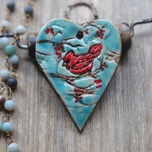 May include: A heart-shaped ceramic pendant featuring a relief-carved red cardinal bird perched on branches with red berries against a light blue glazed background, suspended from a dark metal wire with a strand of small round blue-grey beads.