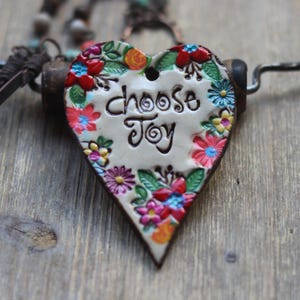 May include: Heart-shaped pendant necklace with the words "Choose Joy" in black script. The pendant is cream-colored and adorned with colorful floral designs in red, blue, yellow, and pink. The necklace has a dark brown leather cord.