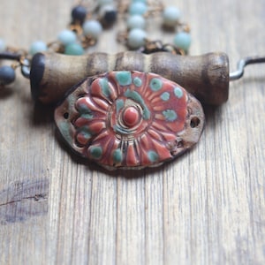 May include: A ceramic pendant featuring a floral relief pattern in coral and teal, attached to a cylindrical wooden bead. The piece is part of a necklace with a dark metal wire frame and a strand of small, round, light blue and grey stone beads.