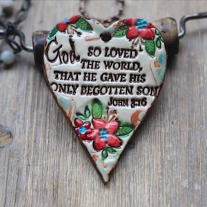 May include: A heart-shaped ceramic pendant featuring a cream-colored glaze with hand-painted red flowers and green leaves. The text reads: God so loved the world, that he gave his only begotten son John 3:16. It hangs from a dark metal chain.