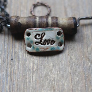 May include: A rustic, handmade corkscrew with a wooden handle and a decorative ceramic tag. The tag is rectangular, with the word "Love" in cursive script, and a speckled turquoise design. A beaded chain is visible in the background.