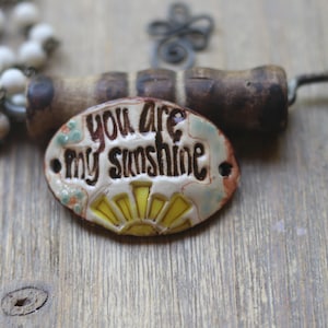 May include: A handmade ceramic pendant necklace with the words "you are my sunshine" in brown lettering. The oval pendant has a yellow sun design and is accented with small floral details. The necklace has a dark brown cord and wooden beads.