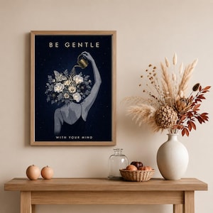 May include: A framed wall art print featuring a dark blue background with a person seen from behind, their head replaced by a bouquet of white and grey flowers and a moth. A hand holds a gold watering can over the flowers. Text reads BE GENTLE WITH YOUR MIND.