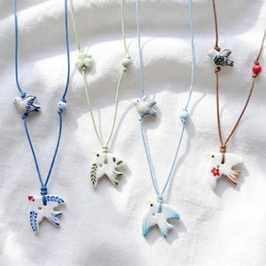 Puede incluir: Una colección de collares con colgantes de pájaros hechos a mano. Cada collar presenta un colgante de pájaro de cerámica blanca con detalles azules, verdes o rojos, ensartado en un cordón. Los cordones son de varios colores, incluyendo azul y marrón.