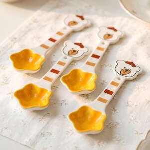 Peut inclure: Quatre cuillères en céramique blanche avec un motif de tête d'ours et des bols en forme d'étoile. Les cuillères ont des rayures orange et marron sur les manches et des bols jaunes. Les têtes d'ours ont des yeux bruns et un petit chapeau orange.