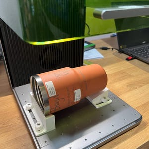 Könnte beinhalten: Ein orangefarbener Becher mit eingraviertem Text, gehalten in einer weißen Halterung, in einer Lasergravurmaschine. Die Maschine hat eine schwarze Basis und eine grüne Oberseite. Der Becher trägt den Namen "Boyd" und weiteren Text.