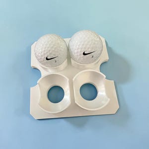 May include: A white plastic golf ball holder with two golf balls. Each ball has a black Nike swoosh and the number 3. The holder has two circular openings and two ball holders.