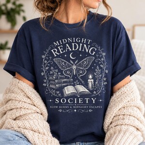 May include: A navy blue t-shirt featuring a white graphic print of a moth, a stack of books, an open book, and a lit candle. The text reads MIDNIGHT READING SOCIETY and SLOW BURNS & MIDNIGHT ESCAPES, surrounded by a delicate vine and star border design.