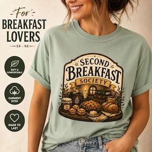 以下が含まれることがあります： セージグリーンのTシャツ。中央には「SECOND BREAKFAST SOCIETY」の文字と、パイ、パン、ジャム、本が並ぶテーブルのイラストが描かれています。デザインにはキャンドルとゴールドブラウンの葉のアクセントが含まれています。