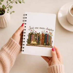 May include: A white spiral-bound notebook featuring a colorful illustration of a row of books on a shelf with flowers and the text Just one more chapter printed above the books. The notebook is held by hands wearing a beige knit sweater sleeve.